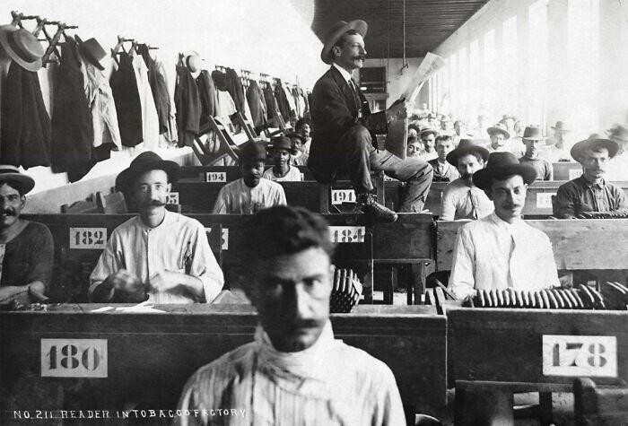 Наемный чтец усердно читает вслух рабочим на кубинской сигарной фабрике (примерно 1900-1910 года). Большинство рабочих были неграмотными, поэтому фабрики нанимали людей, которые зачитывали вслух романы, стихи, научно-популярную литературу и газет